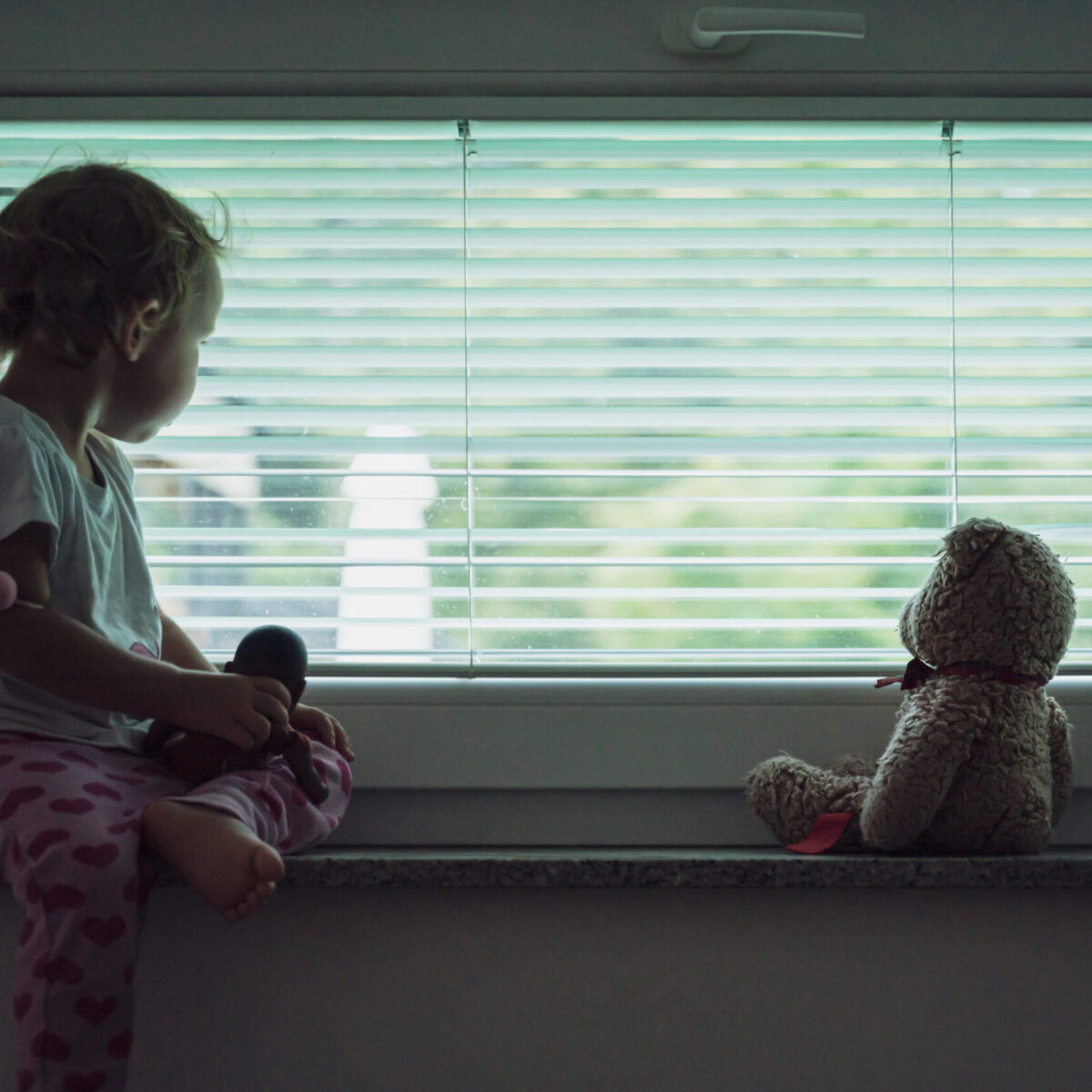 Ein Kind sitzt mit einem Teddybären am Fenster und guckt raus.