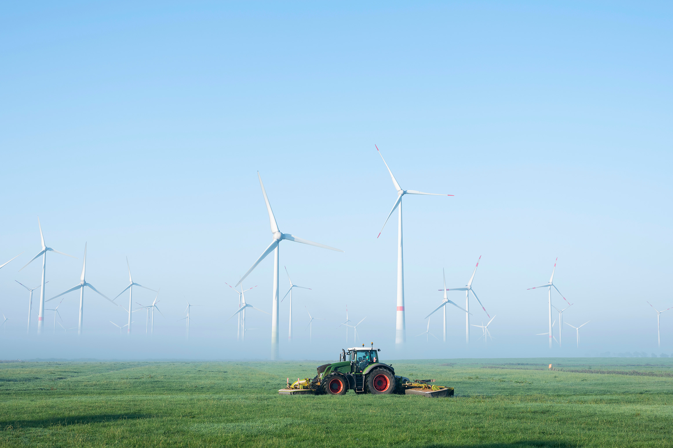 Trecker auf einem Feld mährt Gras. Im Hintergrund stehen im Nebel einige Windräder.
